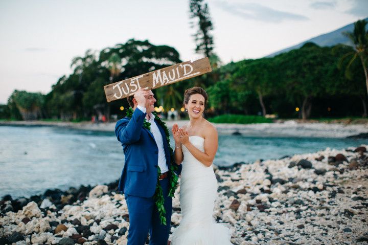 bride and groom standing on beach holding wooden sign that says 