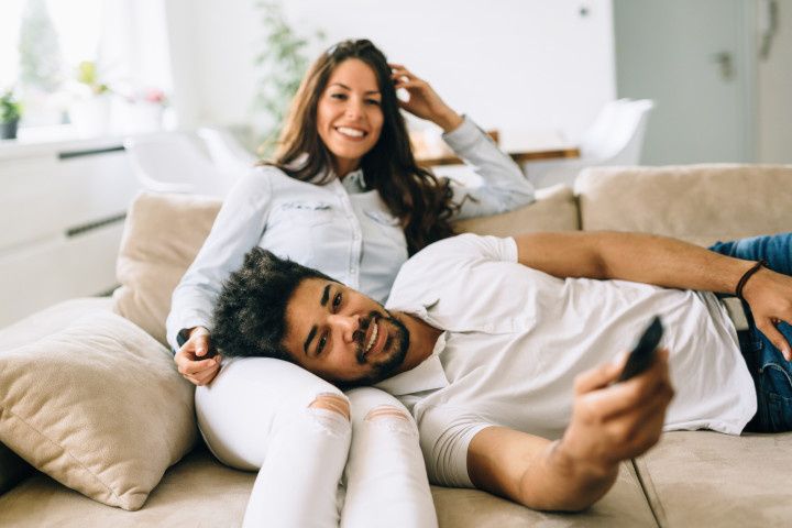 smiling couple sits on couch together man is holding a remote with his head in her lap