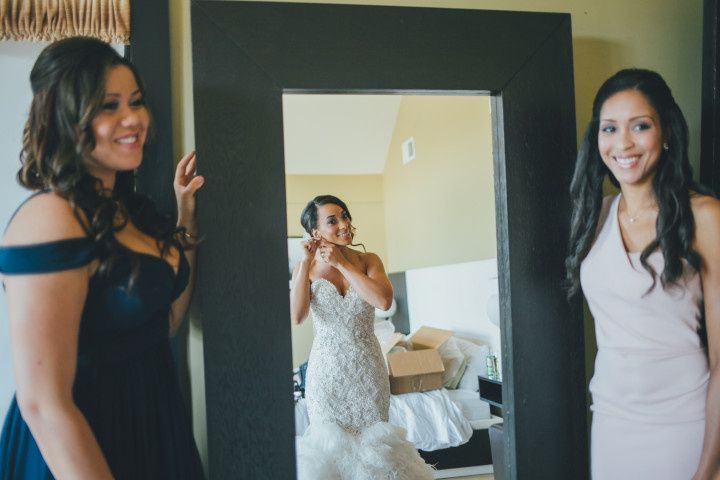 bride looking in mirror while getting ready for her wedding with two bridesmaids watching