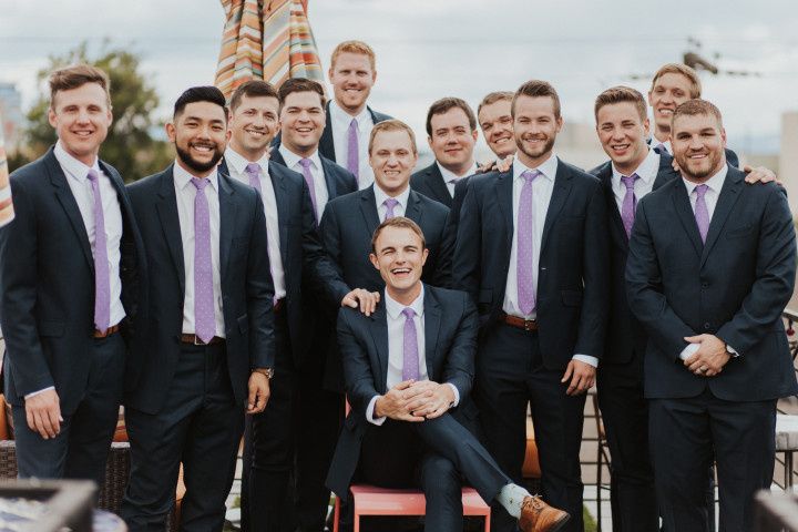 groomsmen wearing navy blue suits and light purple ties stand around groom seated in a chair