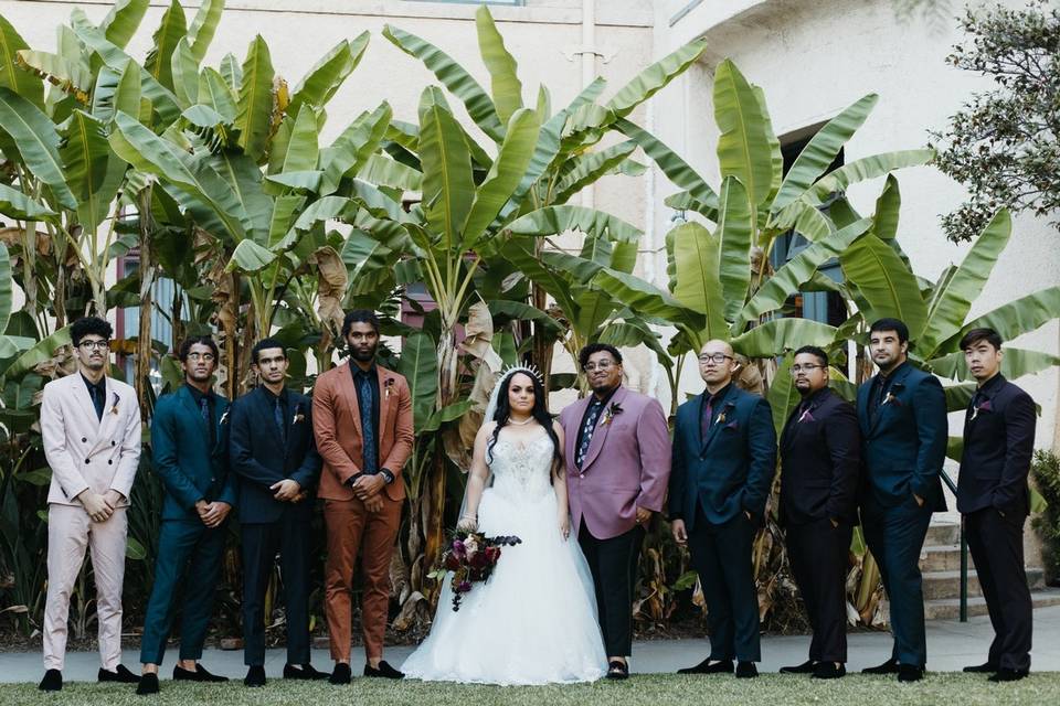 stylish Black couple stands with their wedding party. she is wearing a ball gown with a gold crown and he is wearing a pink suit