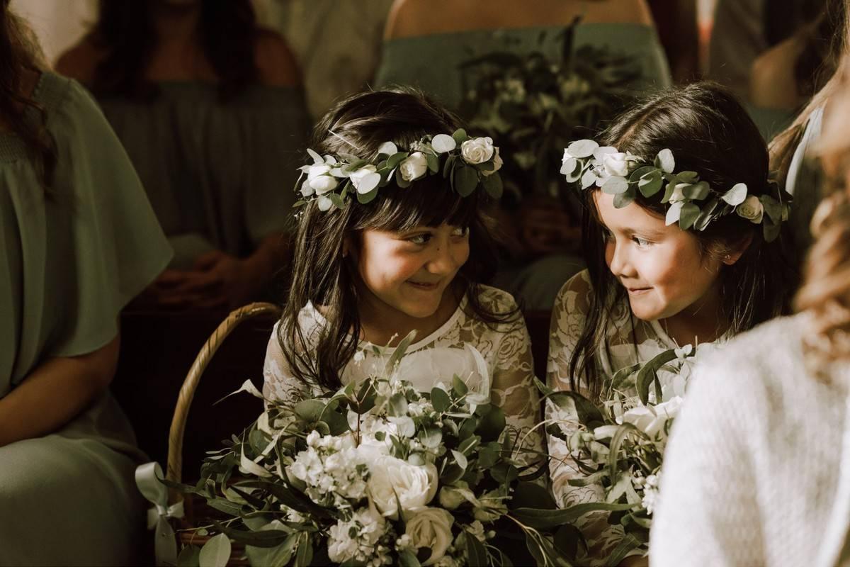 two flower girls wearing greenery crowns sit in chairs during the wedding ceremony and look at each other while giggling