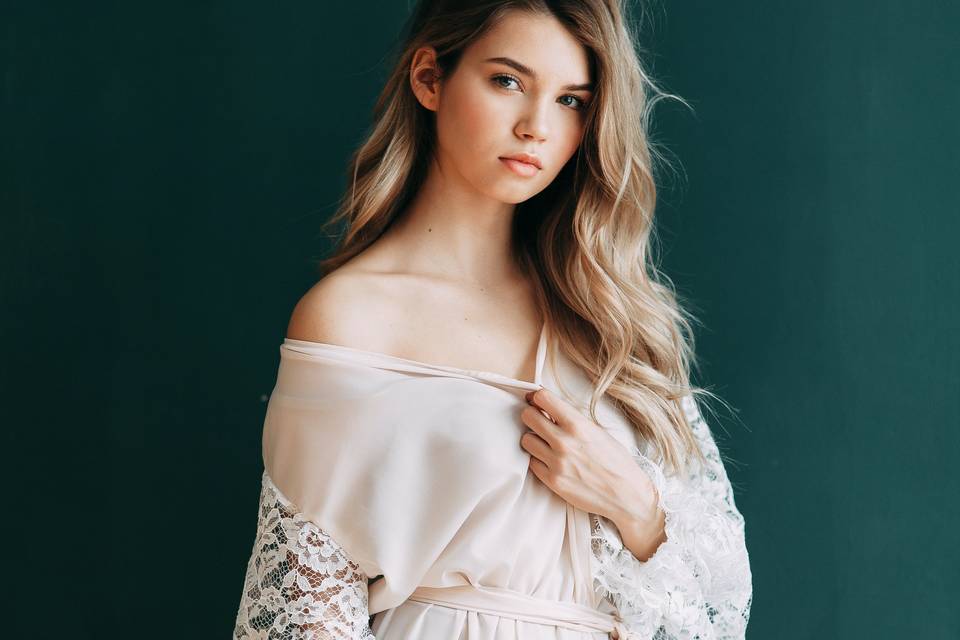 model stands against a plain dark teal wall wearing lace white robe pulled down on one shoulder 