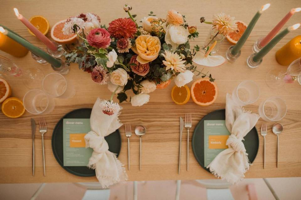 photo of wedding tablescape from above light wooden table with colorful pink, yellow, and orange flower centerpiece with tall colorful taper candles on both sides and fresh orange slices placed throughout the tablescape as decorative detail