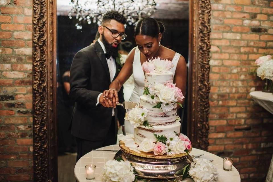 Black bride and groom hold knife together while they cut into a semi-naked wedding cake decorated with white frosting and pink flowers