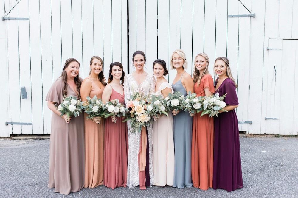 Bride posing with her bridesmaids outside a barn venue all wearing different color dresses