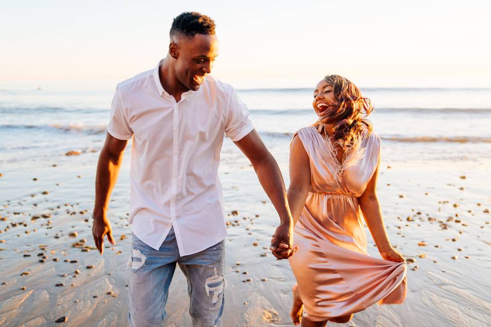 black couple laughs while looking at each other and holding hands walking on a beach at sunset