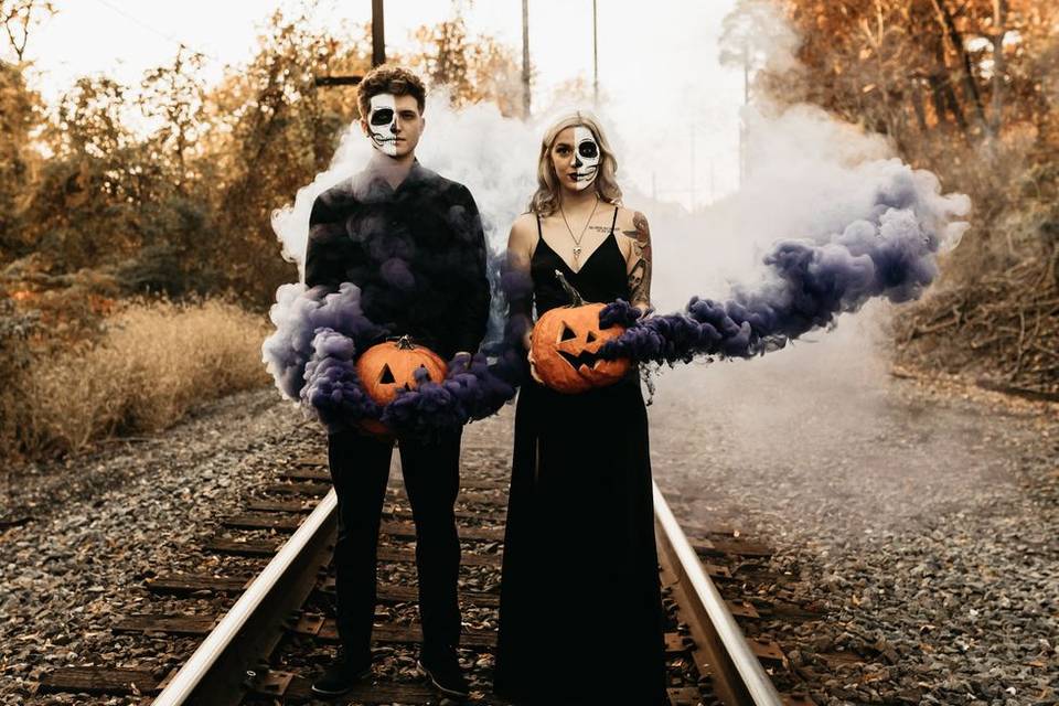 bride and groom with skeleton face paint stand on train tracks holding smoke bomb jack o lanterns