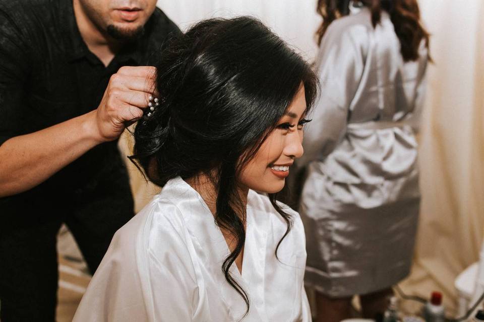 Bride smiles as hairstylist puts the finishing touch on her wedding hairdo, which is a low tousled bun with face framing pieces