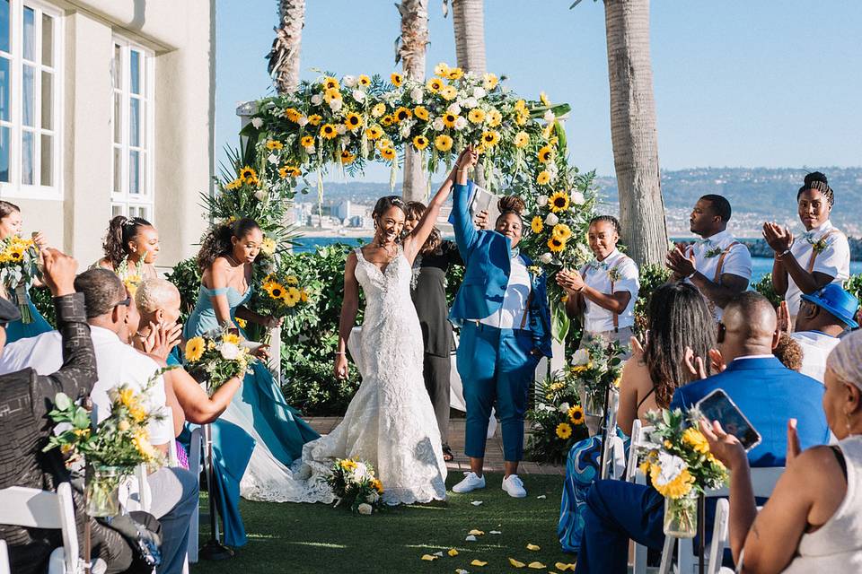 two brides same-sex wedding stand at alter smiling with hands in the air with sunflower ceremony arch and ocean view in the background