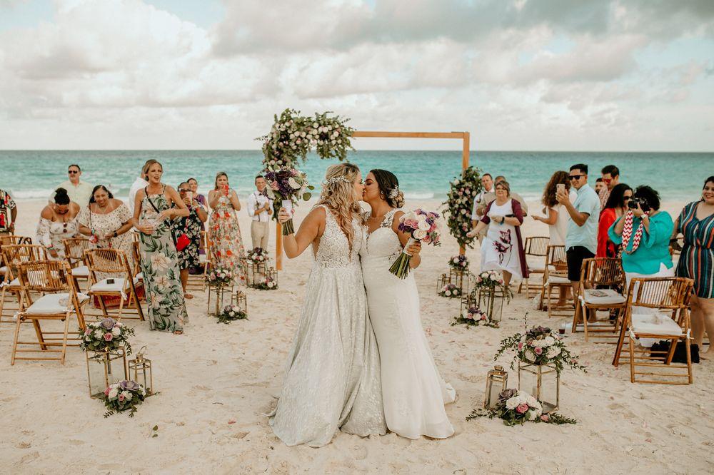 two brides kiss at beach wedding ceremony while guests cheer in the background