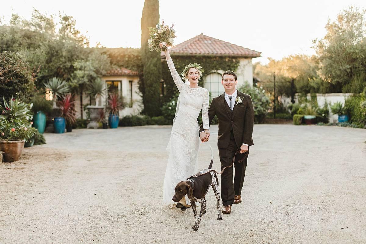 bride and groom walking through gravel courtyard at sunset with their dog bride is raising one arm in the air holding her bouquet and smiling to the camera
