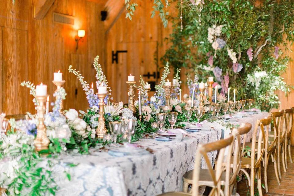 long reception table covered in lace tablecloth and overflowing with light pink, purple, and blue flowers accented with tall gold candle sticks