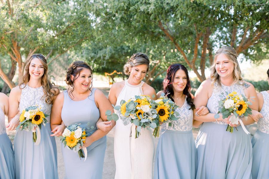 bride links arms with her bridesmaids who are wearing light blue gowns and holding yellow sunflower bouquets