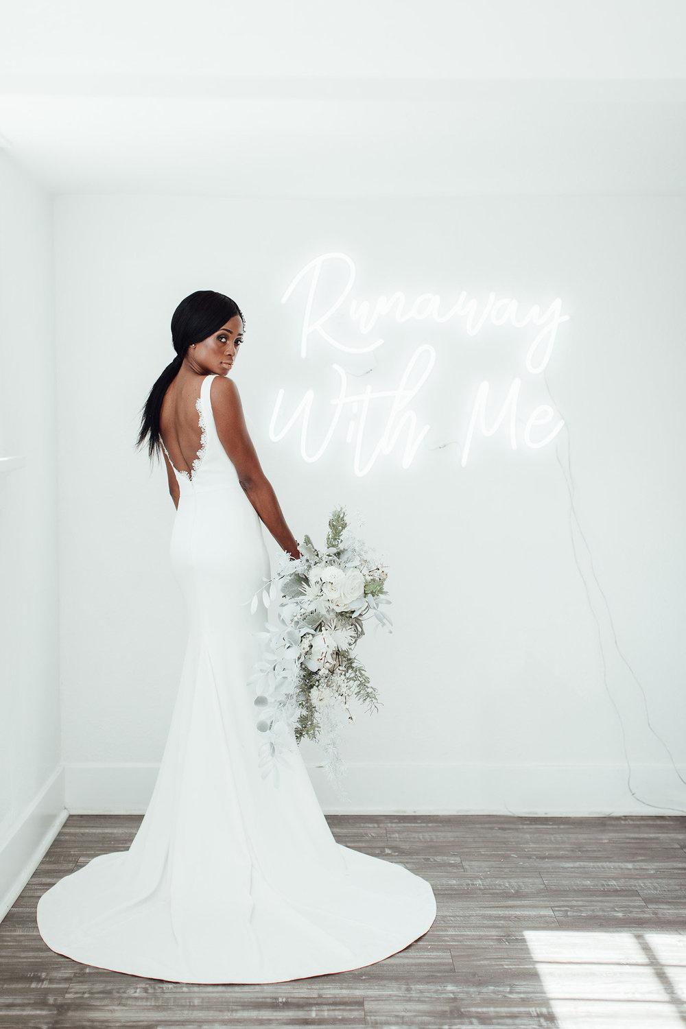 Black bride wears fitted wedding dress and holds a white and green bouquet while standing in front of a neon sign that reads
