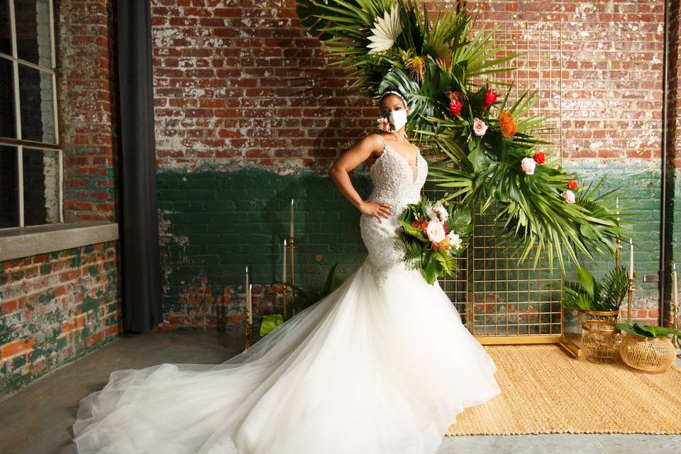Black bride stands in front of tropical floral ceremony backdrop against an indoor brick wall. She is wearing a wedding dress with a long train and a protective face mask decorated with a flower