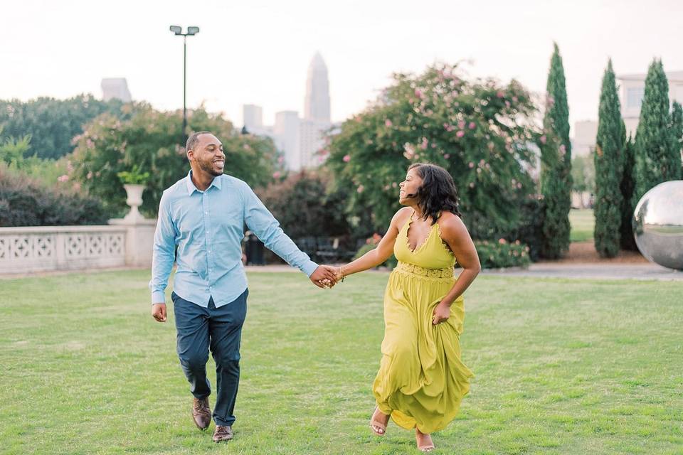 Black couple holds hands as they run through a manicured lawn. She is wearing a long lime green dress and he is wearing a blue button-down shirt