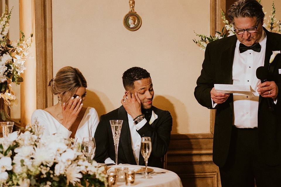 bride and groom wipe tears from their eyes while seated at a table with father giving wedding speech