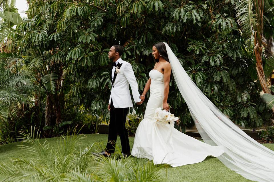 bride wearing cathedral-length veil holds groom's hand as they walk through a garden