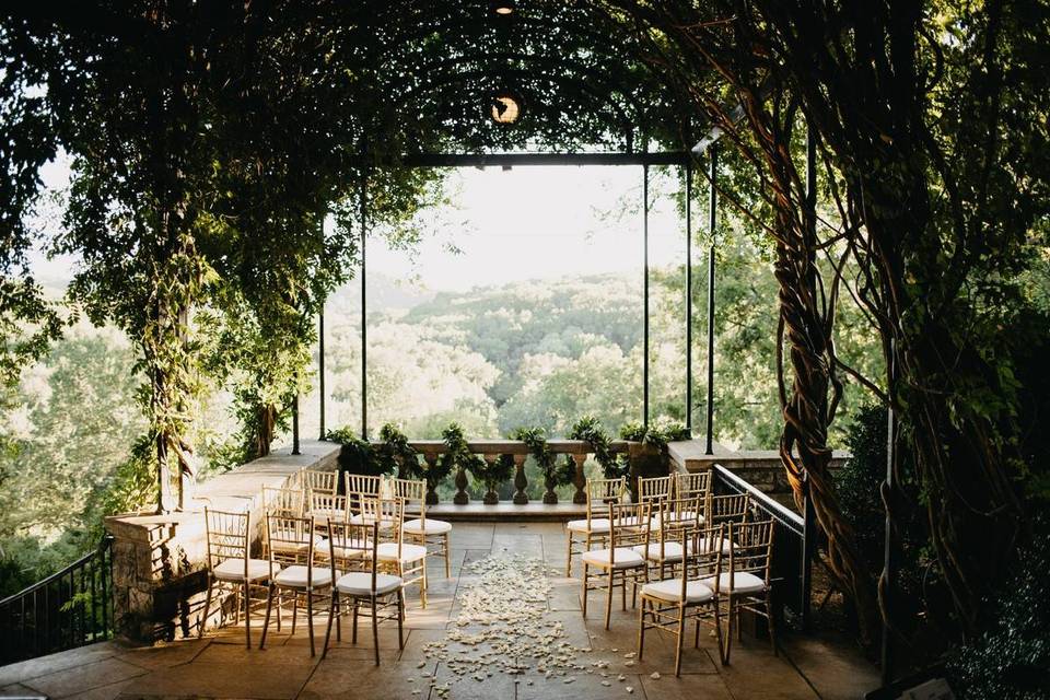 stone terrace under an archway of vines and greenery. rose petals are scattered on the floor and the terrace overlooks hills in the distance