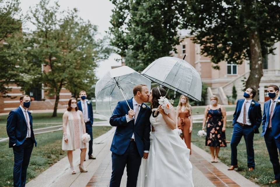 bride and groom kissing under umbrellas with guests wearing masks watching