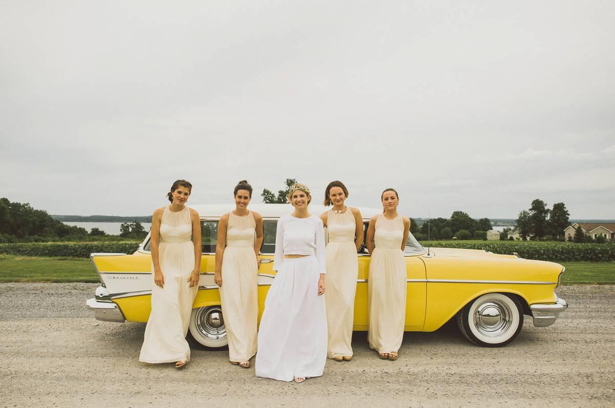 Bride posing with bridesmaids in front of a yellow car wearing buttercream yellow bridesmaid dresses