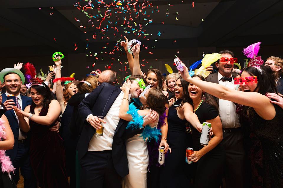 couple kisses on dance floor surrounded by guests wearing new year's hats and glasses while confetti drops from the ceiling