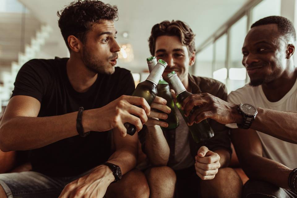 three guys sit on a couch and toast with beer bottles