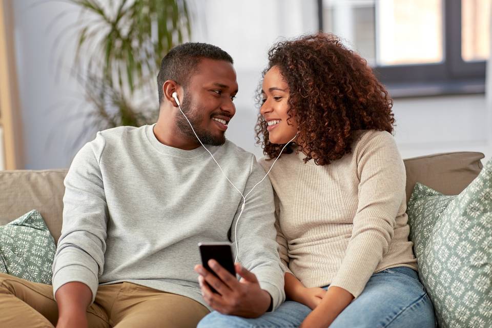 couple sitting next to each other on a couch, each with an ear bud in an ear holding an iphone