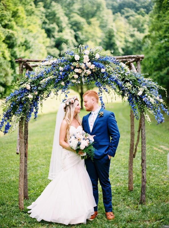 bride and groom smile at each other while standing under handmade wooden arch decorated with blue delphinium