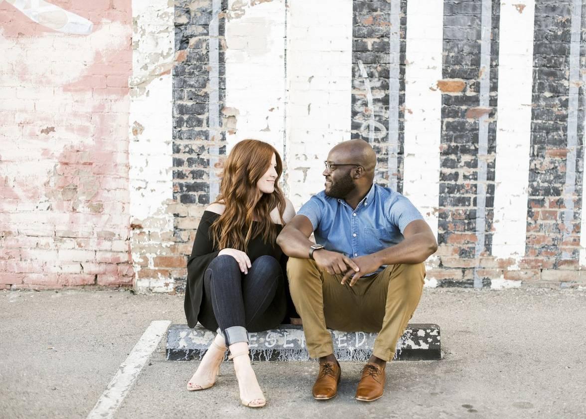 interracial couple sit on the sidewalk and look at each other while holding hands