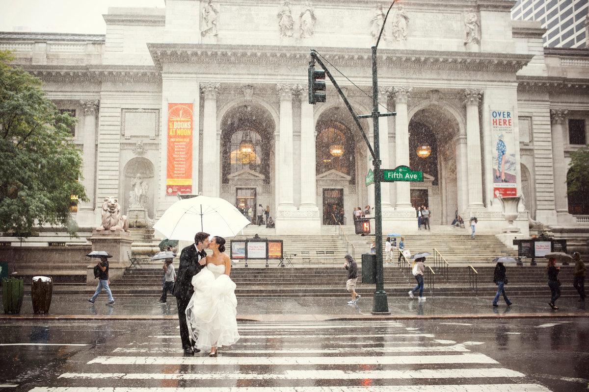 bride and groom cross the street in front of new york public library under an umbrella in the rain