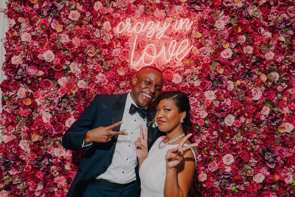groom and bride standing in front of flower wall with crazy in love neon sign