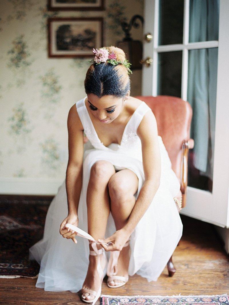 Black bride is seated while fastening ankle straps on her high heels with fresh flowers adorning her sleek bun hairstyle