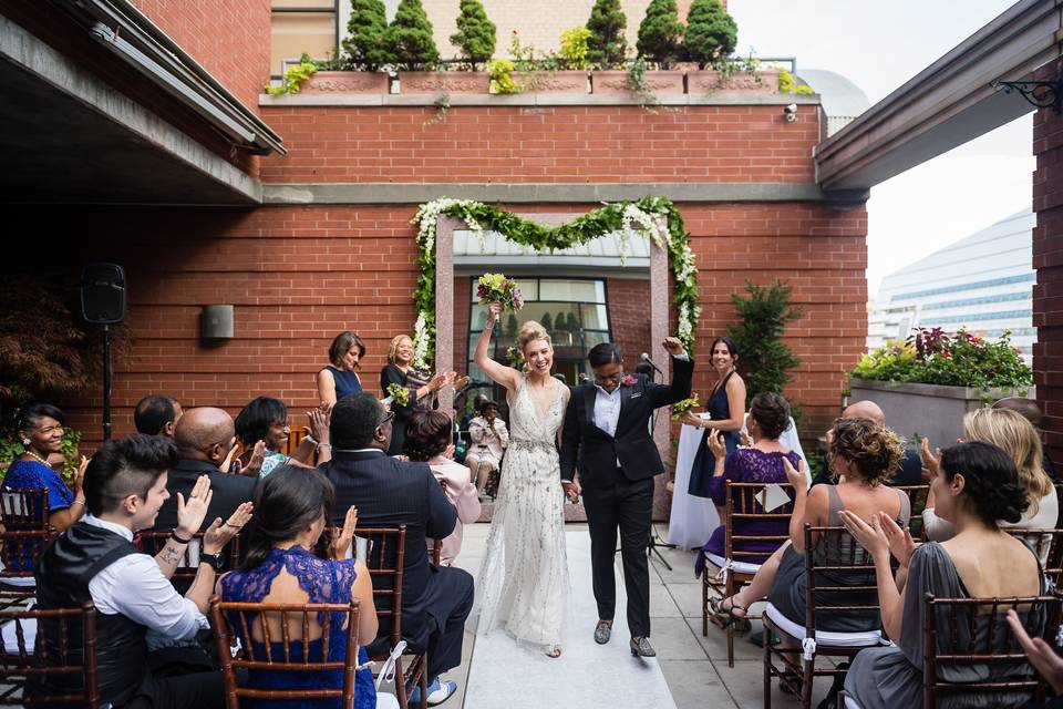 LGBTQ+ interracial couple celebrates as they walk down the aisle at outdoor rooftop venue in NYC after exchanging their wedding vows