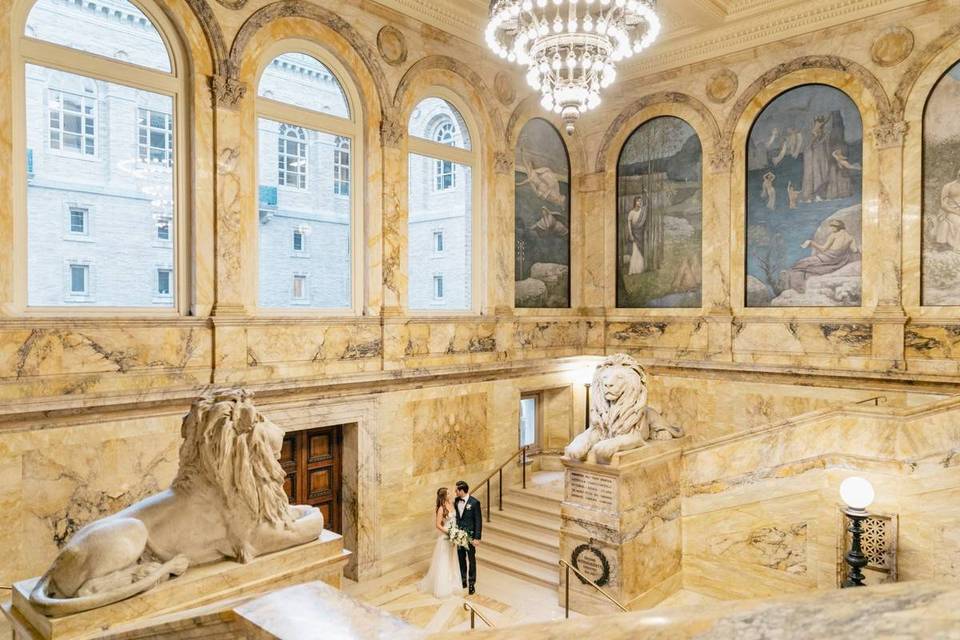 bride and groom stand on the marble staircase next to lion statues at boston public library