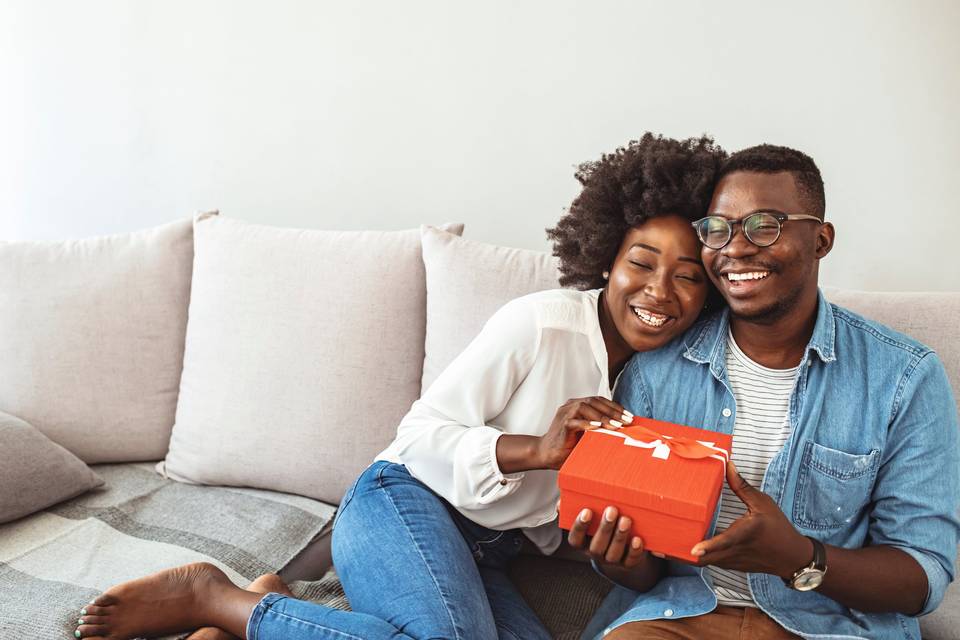 couple sitting on couch holding a red gift box and laughing