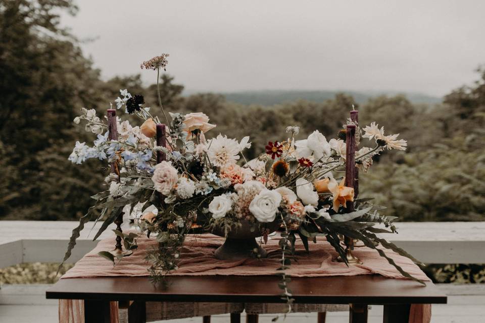 boho wedding centerpiece with white, blush, blue, and dark orange flowers on a table outdoors with tall burgundy candles on both sides