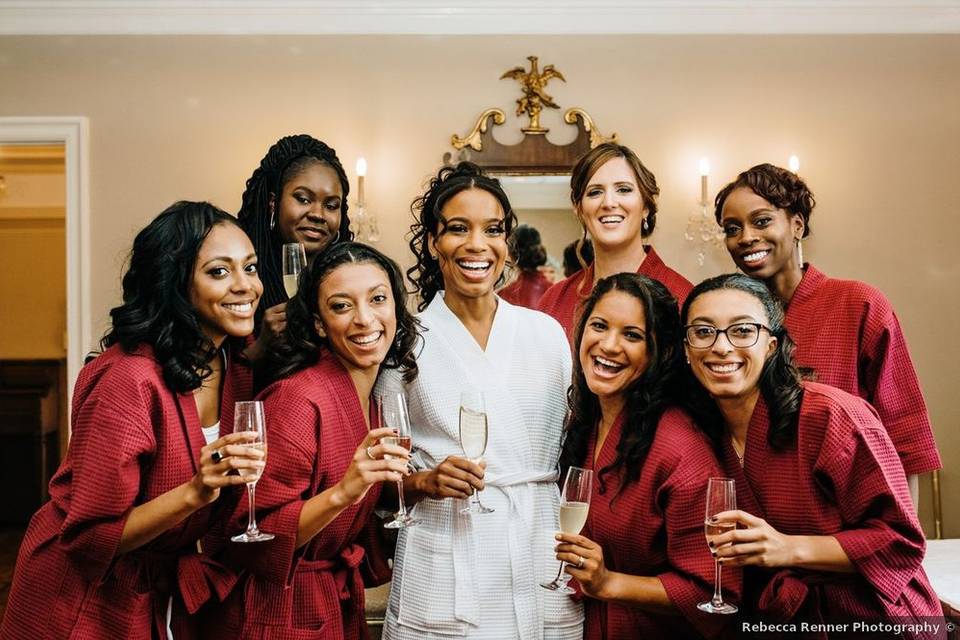 Black bride wears a white spa robe and is surrounded by her group of bridesmaids who are wearing burgundy robes. They are holding glasses of champagne in their hands 