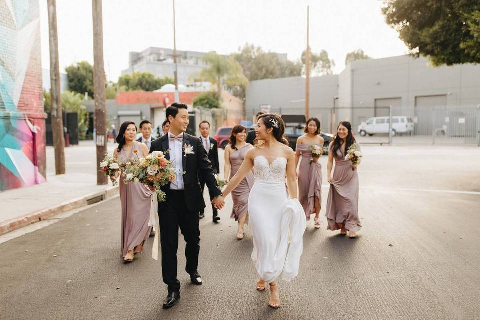 asian couple holding hands looking at each other and smiling walking down the street with wedding party behind them