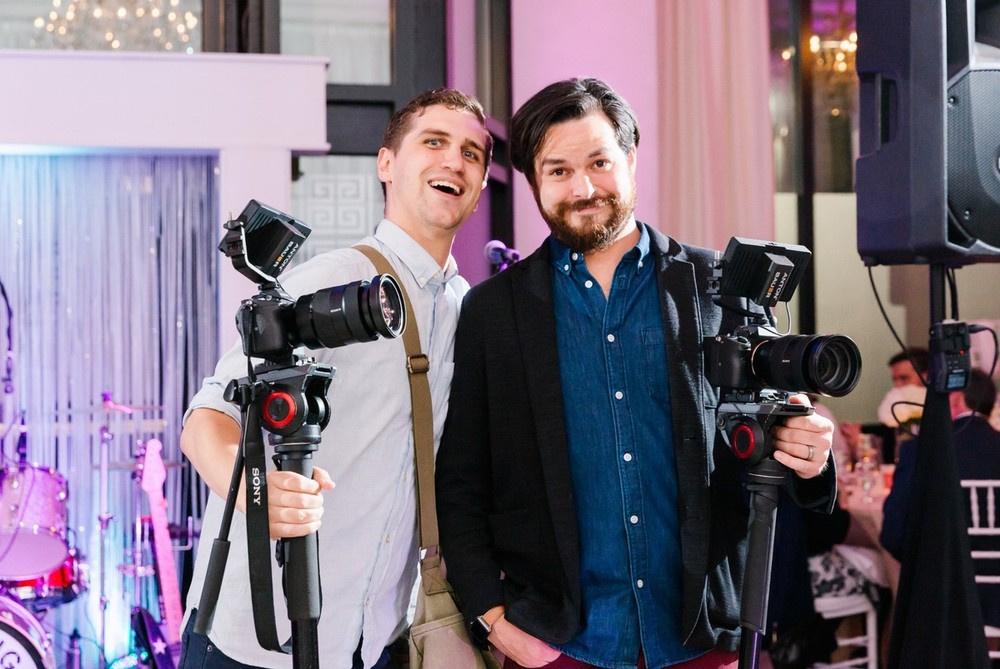 team of two male videographers pose for a photo at wedding reception with their equipment