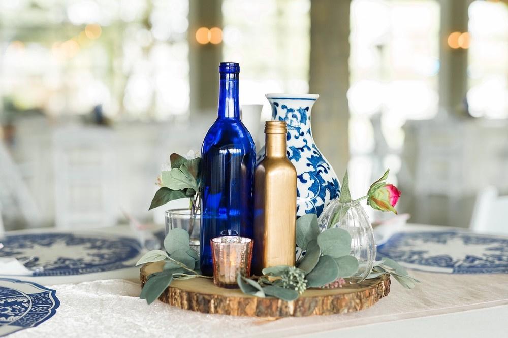 rustic wedding centerpiece with mismatched blue and gold glass bottles, greenery, and votive candles on a hand-cut wooden slab