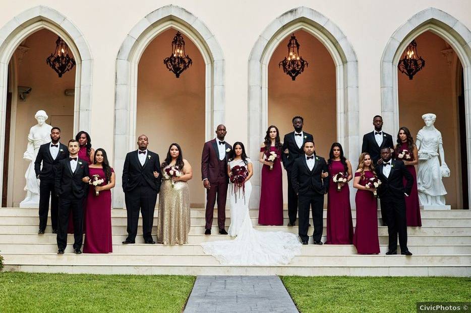 bride and groom, a black couple, stand on the steps of a museum with their wedding party who are wearing tuxedos and long burgundy gowns