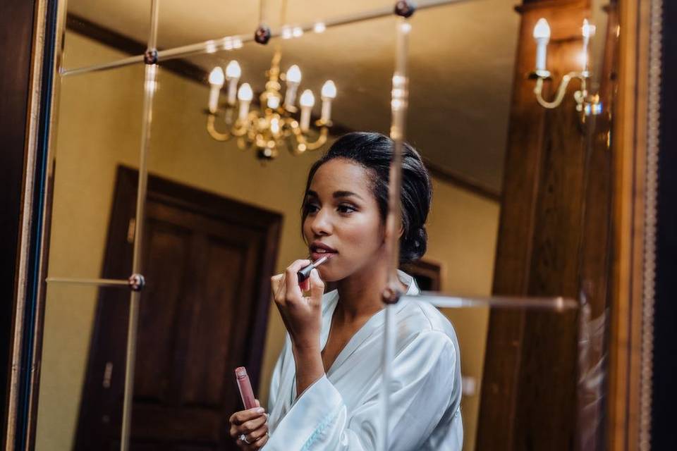 Black bride stands in front of the mirror and applies pink lip gloss. her hair is styled into a low bun