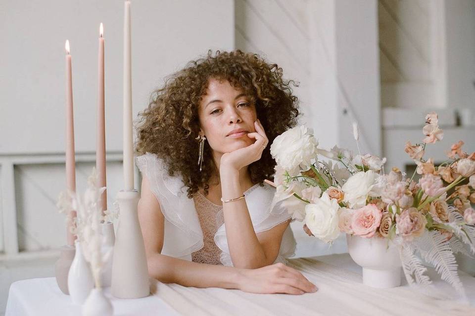 Bride sits at a table and looks into the camera with her chin in her hand and blush wedding centerpiece on one side with long taper candles on the other side