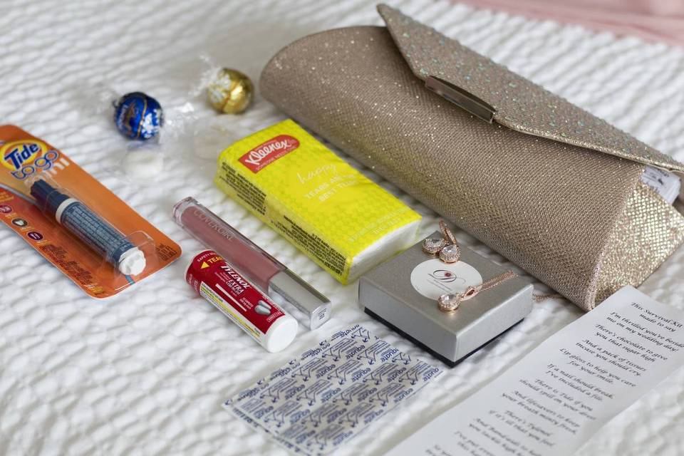 gold clutch with various items laid on a bed tissues, tide pen, tylenol, chocolates