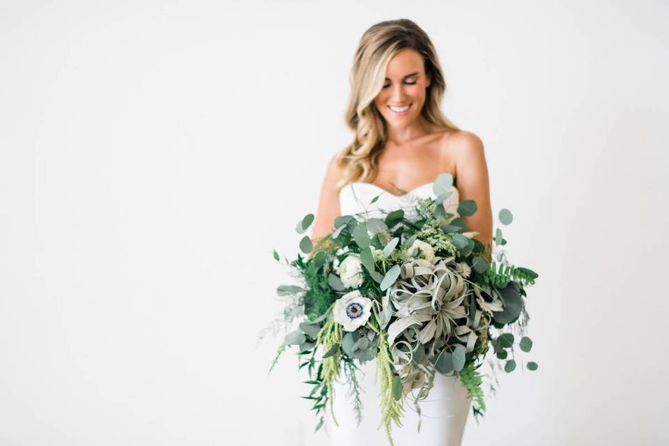 bride stands against white wall holding beach wedding bouquet of greenery eucalyptus anemones and air plants