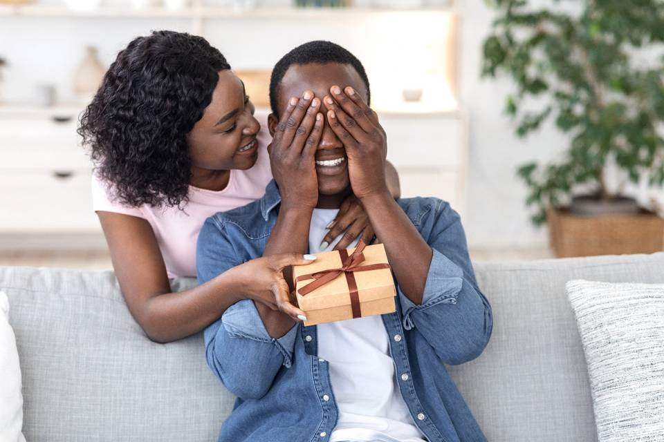 Boyfriend sitting on sofa covering eyes with eyes as girlfriend presents a gift from behind him