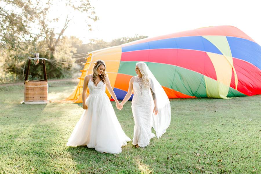 two brides hold hands as they walk in front of a hot air balloon on the grass