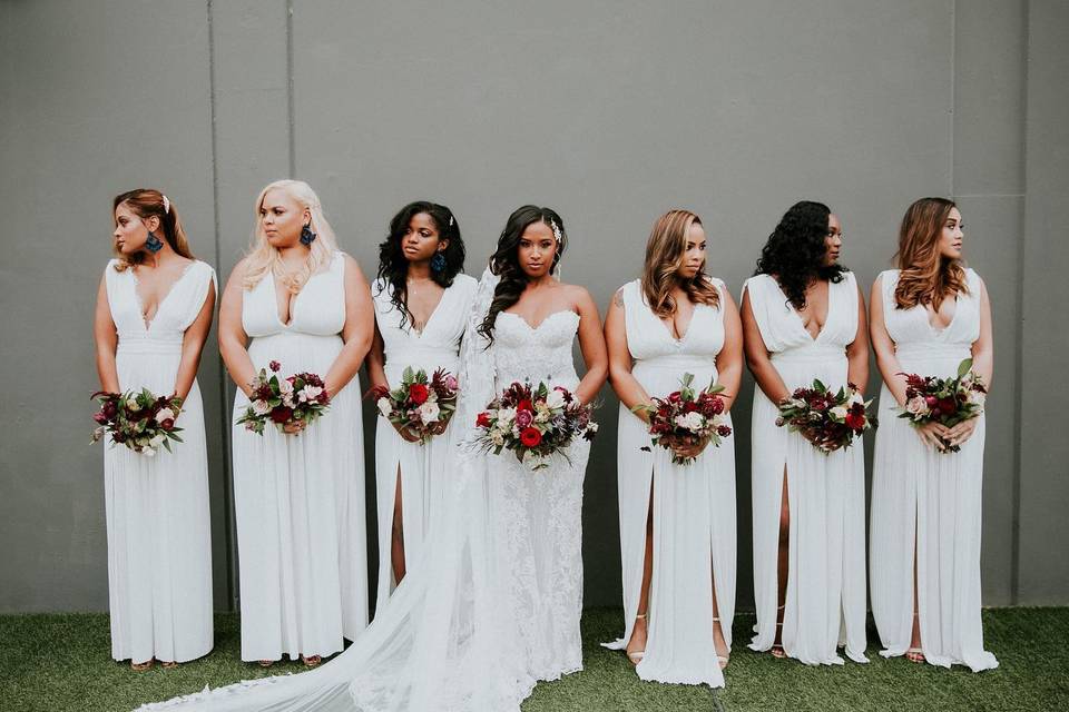 Black bride stands and looks into the camera while holding bouquet with bridesmaids standing on both sides wearing white floor-length dresses